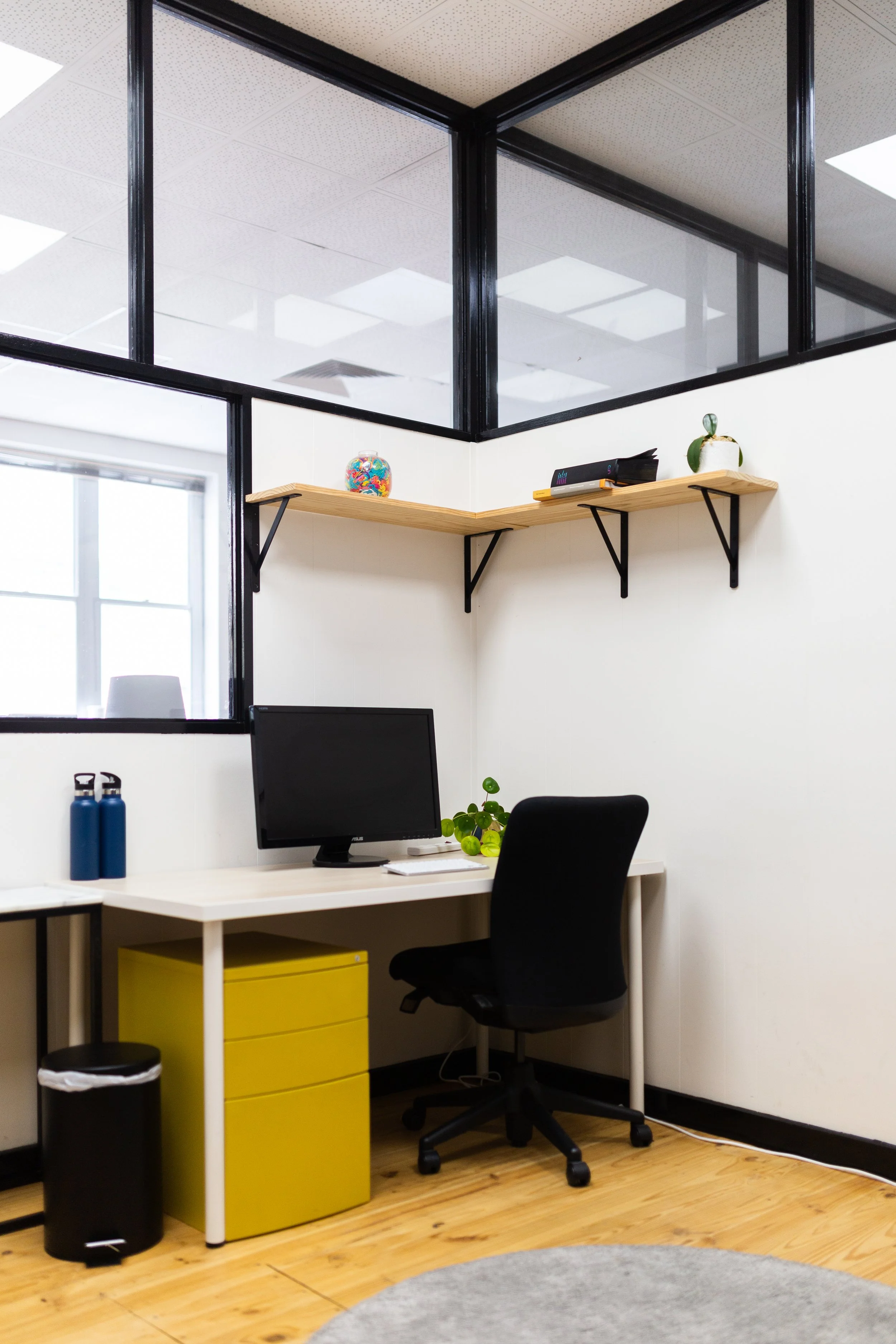 Office workspace with a white desk, black office chair, computer monitor, green plant, wooden shelf with decorative items, two blue water bottles, and a yellow filing cabinet.