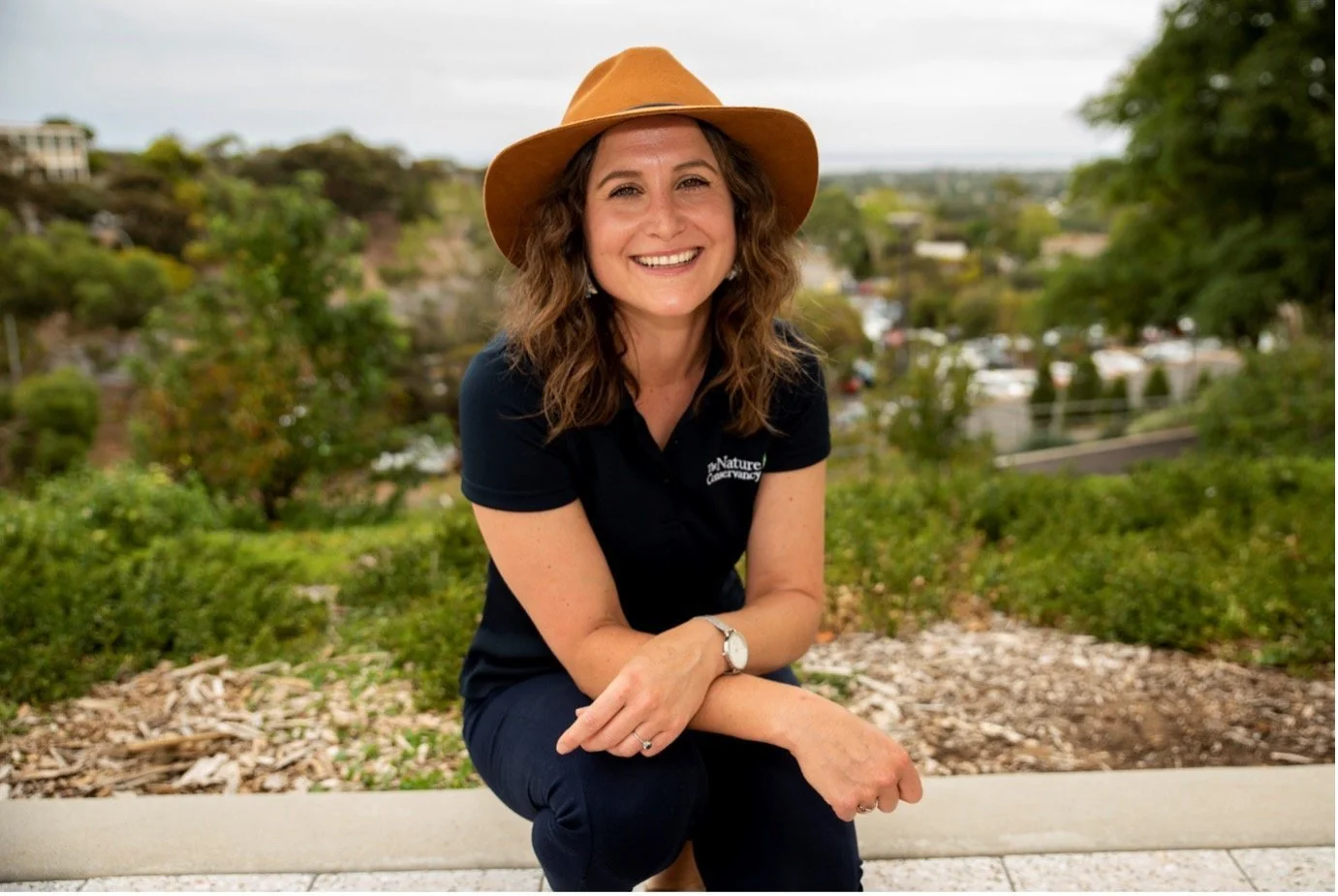 Smiling woman with wavy brown hair wearing a tan hat, black polo shirt, and dark pants, sitting outdoors with a scenic view of trees, buildings, and a cloudy sky in the background.