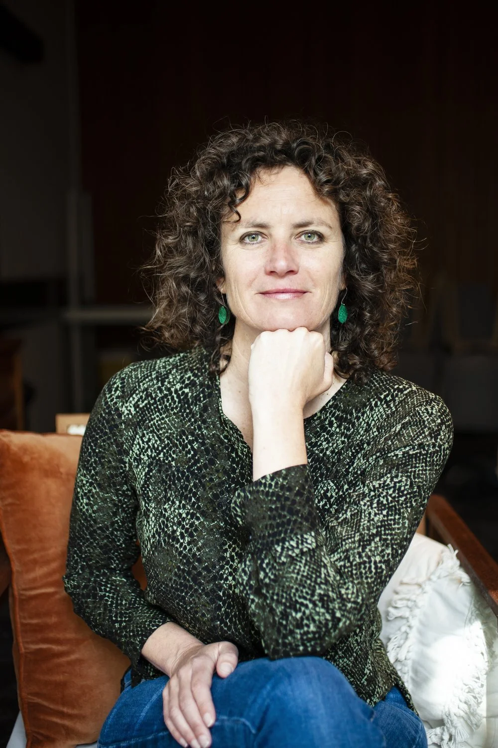 Portrait of a woman with curly dark hair, green earrings, sitting on an orange sofa with a green patterned top, resting her chin on her hand, in a room with brown walls.