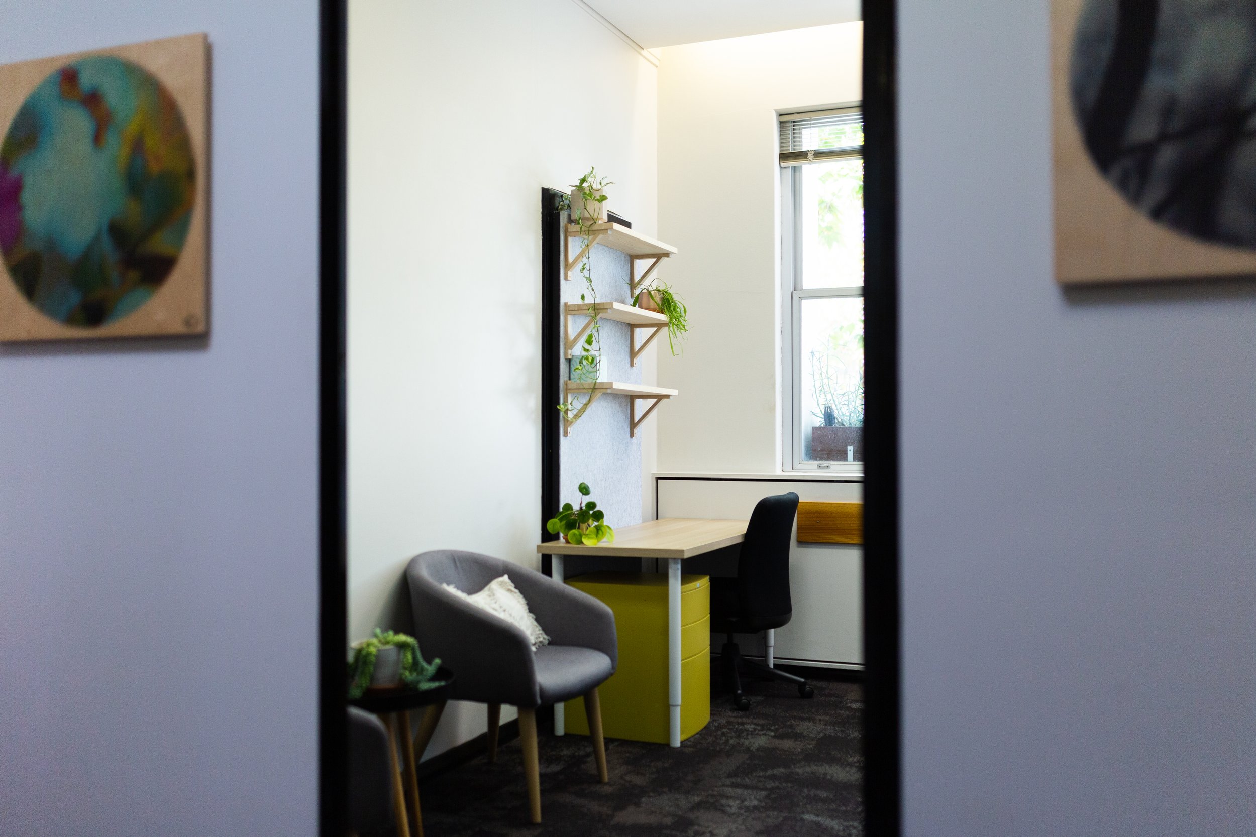 Small office with a desk near a window, gray armchair, and wall shelves with plants, viewed through an open doorway.