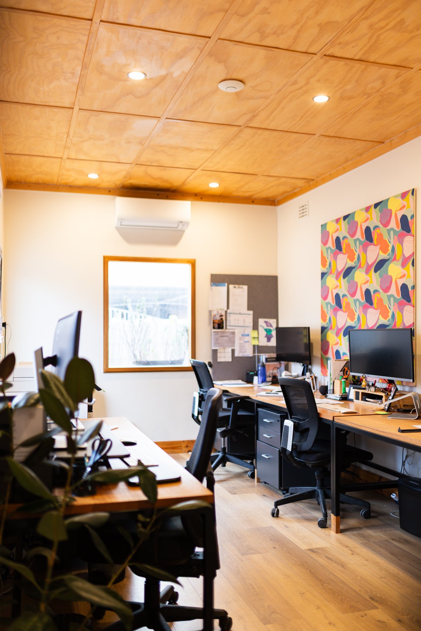 Office room with wooden ceiling, multiple work desks with computers, chairs, a window, motivational bulletin board, and colorful abstract artwork on the wall.