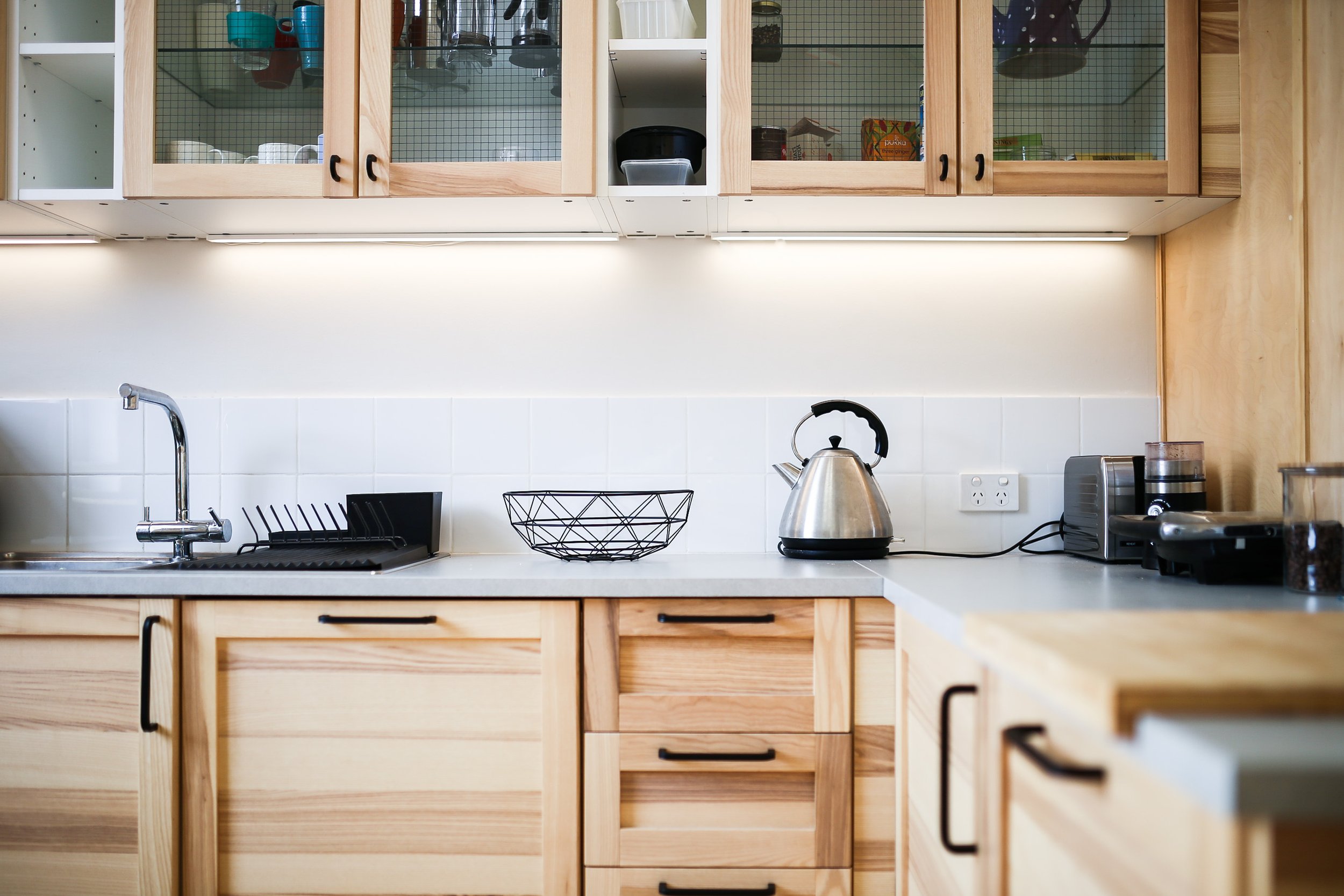 Kitchen with light wood cabinets, white tile backsplash, stainless steel kettle, black dish rack, white countertop, and small appliances.
