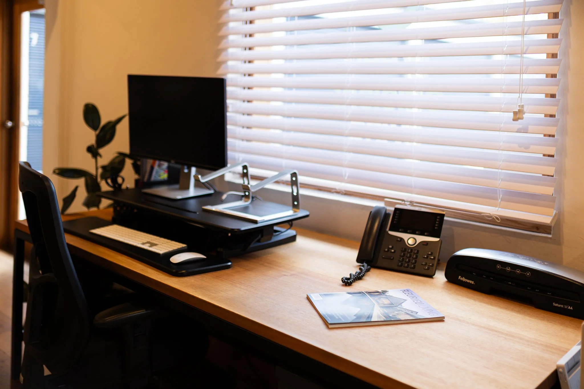 Office desk with a computer monitor, keyboard, mouse, phone, and office supplies, next to a window with blinds.