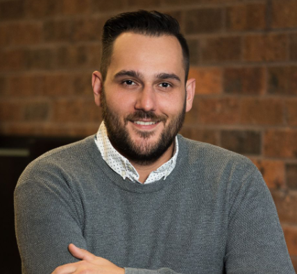 A man with dark hair and a beard, smiling, wearing a gray sweater over a white collared shirt, standing in front of a brick wall.