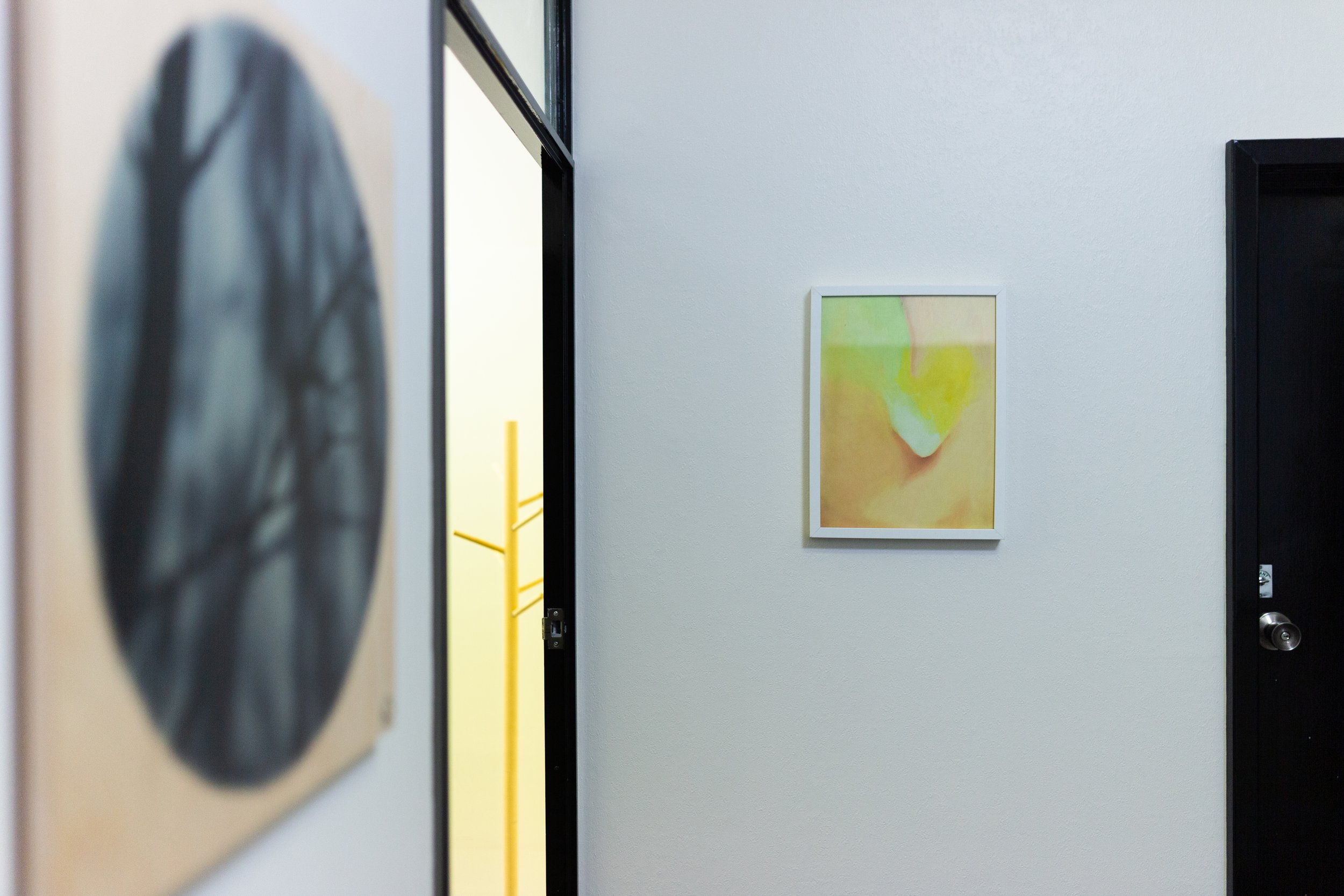 Hallway with a white wall, a framed abstract painting, a black door, and part of a yellow coat rack visible in the background.