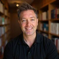 A man with short, dark hair and a beard smiling, standing in front of bookshelves in a library or bookstore.