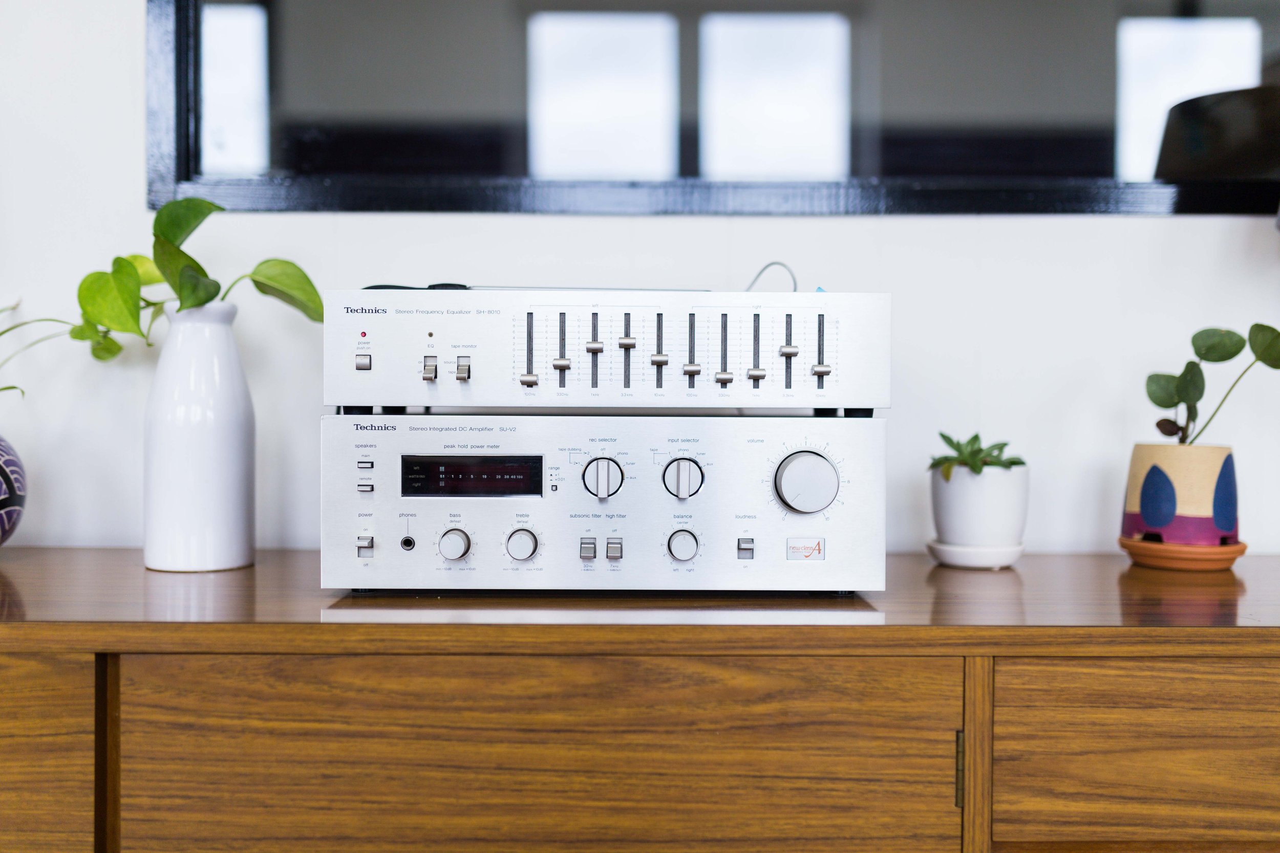 A vintage white stereo system with a tuner and amplifier placed on a wooden sideboard, flanked by various potted plants, with a TV screen in the background.