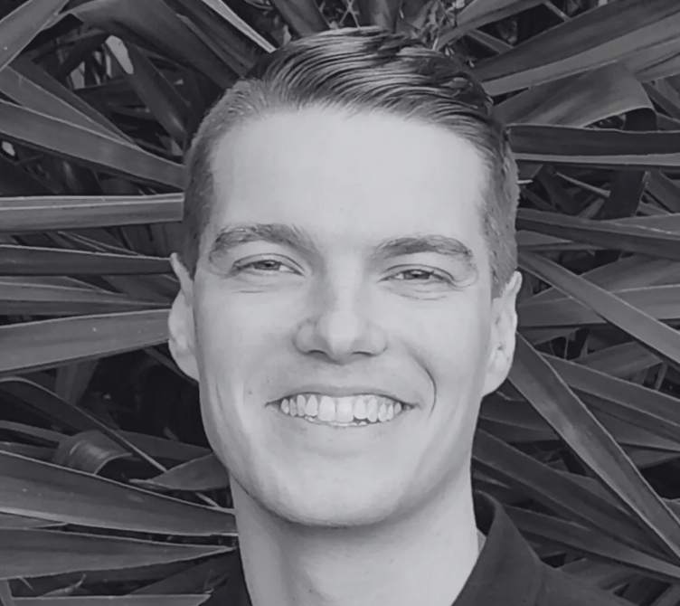 A young man with neatly combed hair smiling in front of large, spiky leaves in a black and white photo.