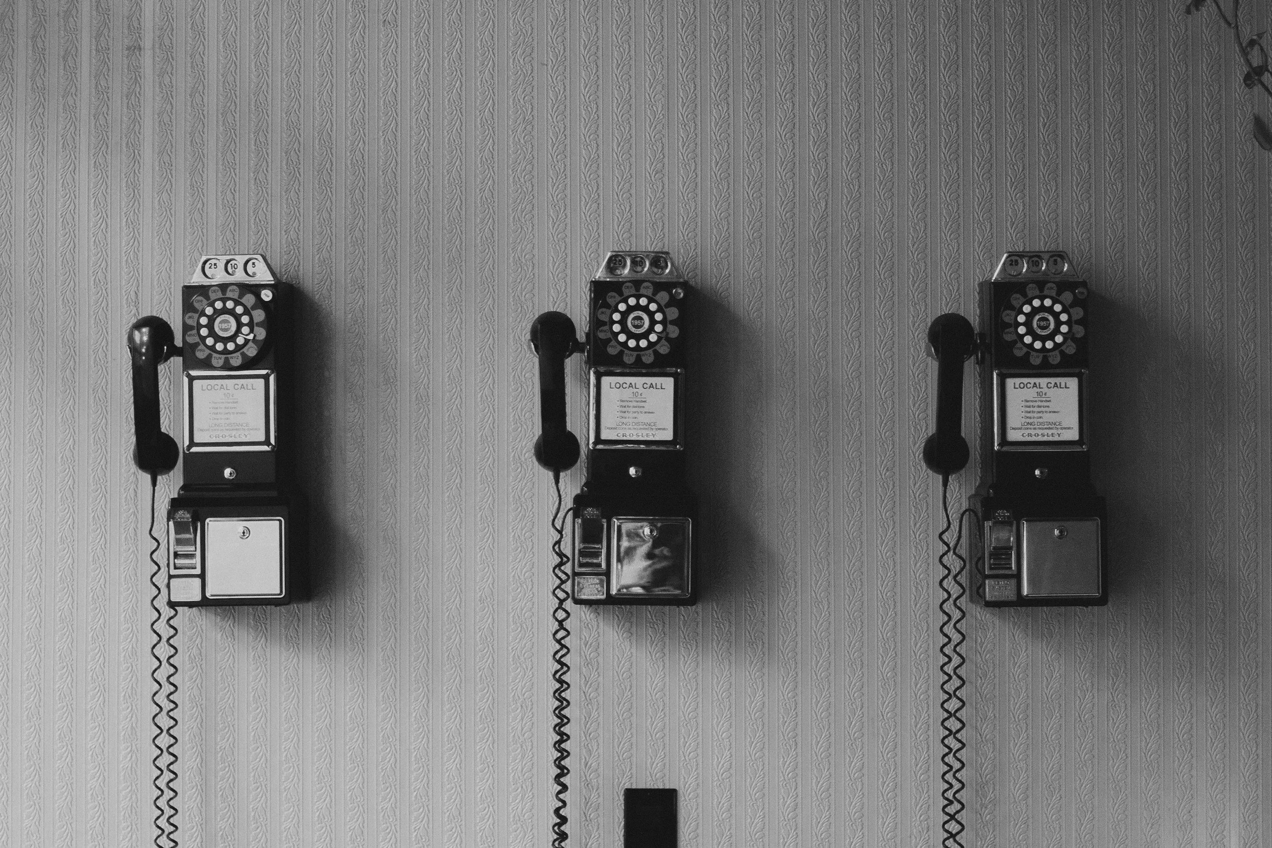 Three vintage rotary dial payphones mounted on a wall with patterned wallpaper.