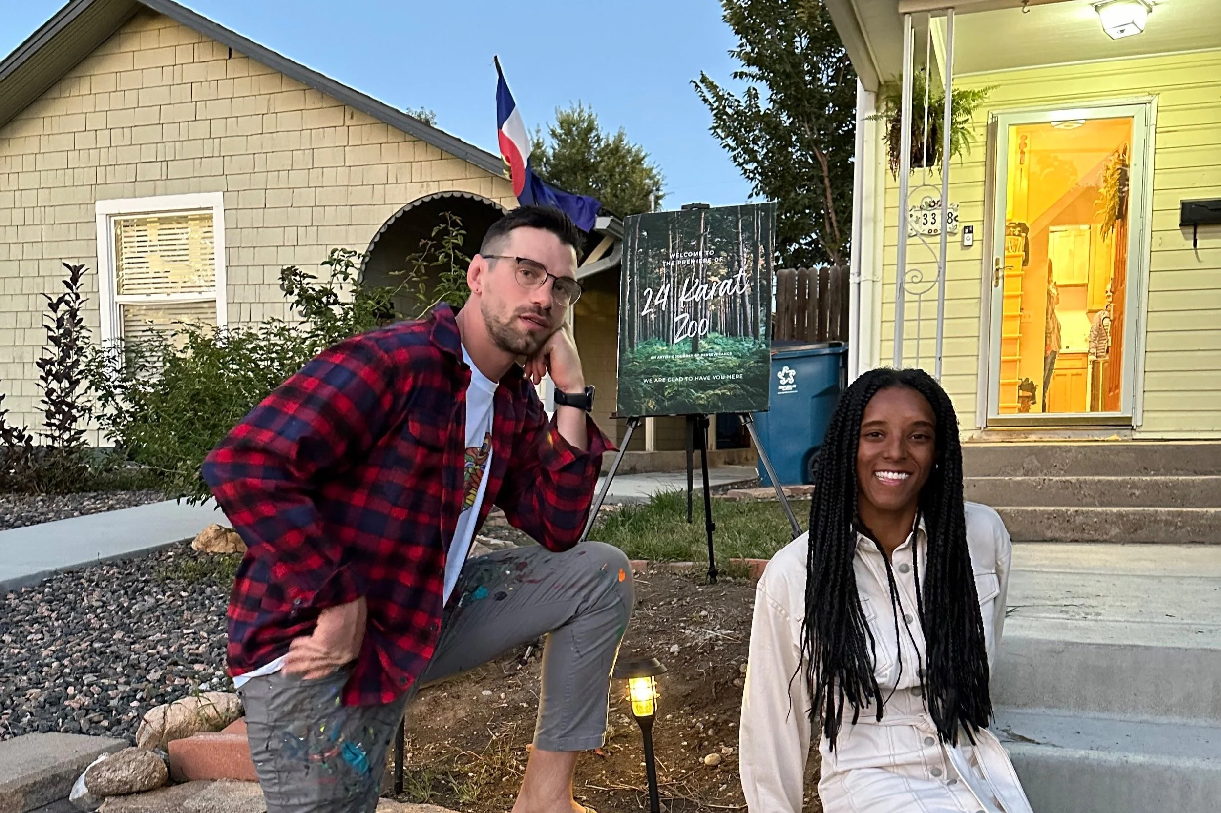 A man and a woman are pictured outside a house during evening time. The man is kneeling on one knee, wearing glasses, a red plaid shirt, and gray pants with paint stains, resting his chin on his hand. The woman is sitting on the steps, wearing a white outfit and smiling. Behind them, a sign reads '24 Karat Zoo,' and the house has a yellow exterior with an illuminated interior visible through the open door.