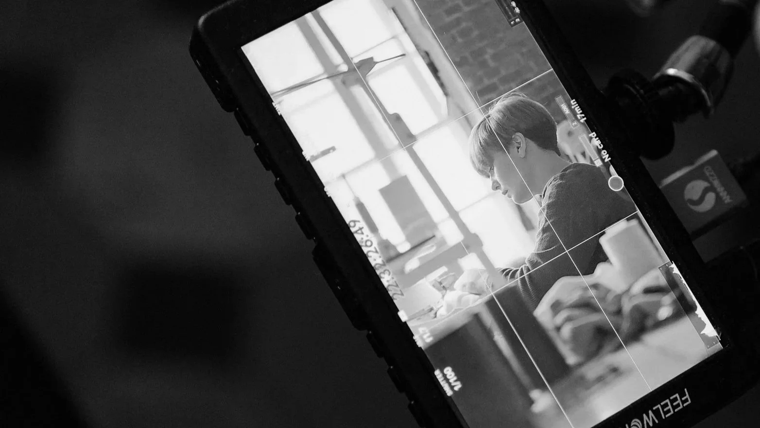 A black and white photo of a mirrorless camera for social media content creation display showing a woman with short hair sitting at a table, writing or working, in a brightly lit room with large windows.