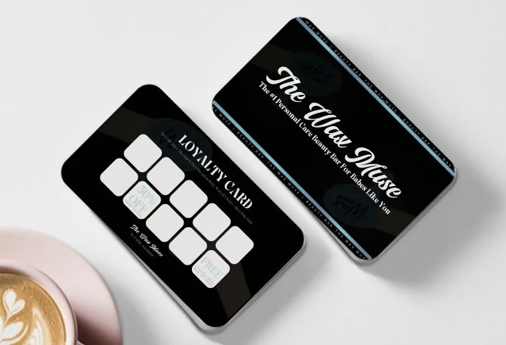 Black loyalty card and a black cardholder with the text "The Wax Muse" on a white surface, next to a pink cup of coffee with latte art.