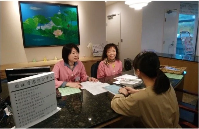 Two women in pink uniforms sitting at a reception desk across from a woman in a tan jacket in an office or clinic setting, with a large television or monitor displaying a nature scene on the wall behind them.