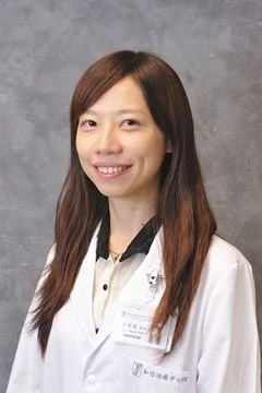 A woman wearing a white lab coat, smiling, with long brown hair, standing against a gray background.
