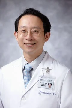 Portrait of a smiling male doctor in a white coat, wearing glasses, with an identification badge, against a gray background.