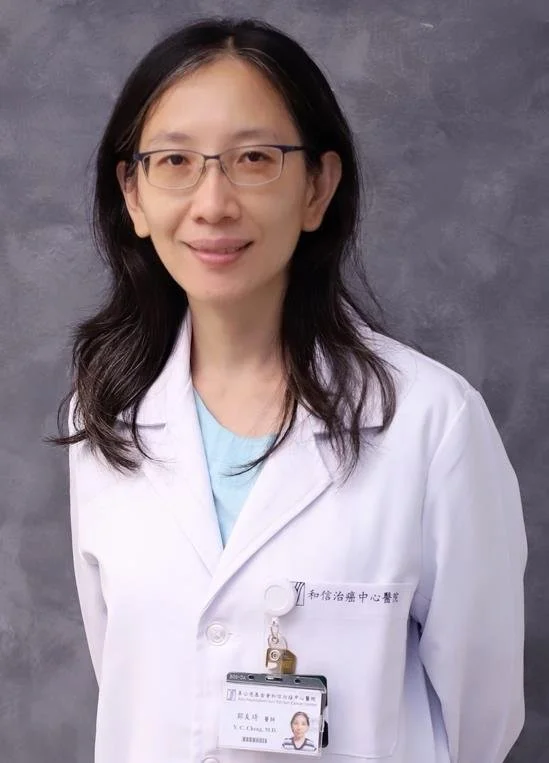 A woman with long dark hair, wearing glasses, a white lab coat, and a light blue shirt, smiling, standing against a grey background.