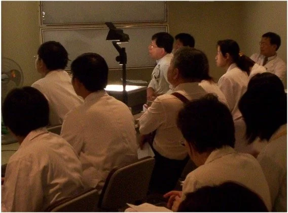 A group of people in white lab coats attending a lecture or presentation in a classroom or conference room.