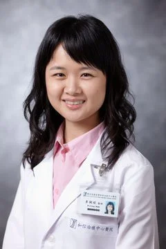 A young Asian woman in a white medical coat with a name tag, smiling against a gray background.