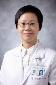 A woman in a white medical coat with glasses, short black hair, and a name badge, standing against a gray background.