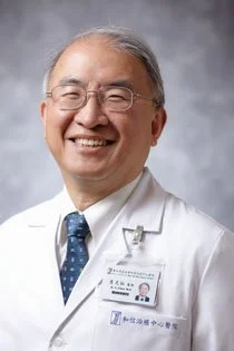 Smiling middle-aged Asian male doctor in white coat with glasses, gray hair, and a name badge, standing against a gray background.