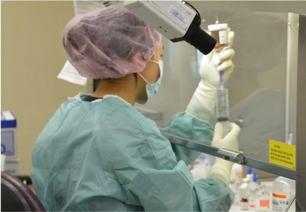 A scientist in a lab coat, wearing a pink surgical cap, face mask, and gloves, holding a pipette in a laboratory with scientific equipment and supplies.