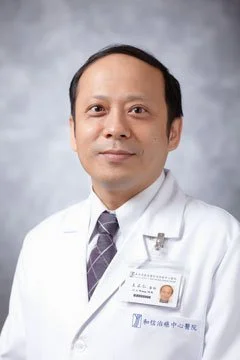 Portrait of a male doctor wearing a white coat and a name badge, against a gray background.