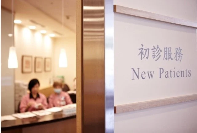 A hospital or clinic waiting area with staff in pink uniforms behind a reception desk, and a sign indicating 'New Patients' in both Chinese and English.