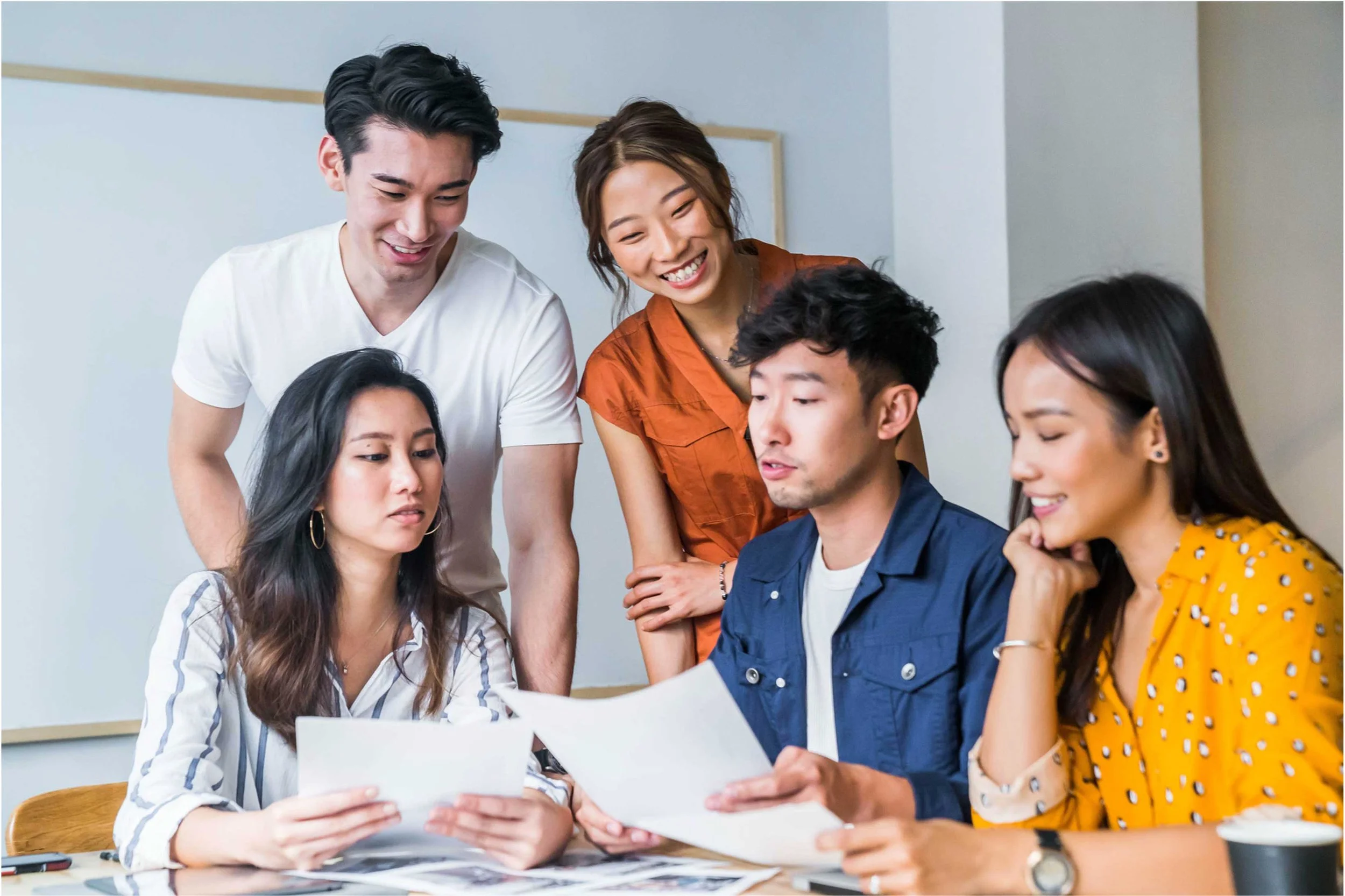 Group of five young adults collaborating around a table, reviewing papers and discussing, in a modern office setting.