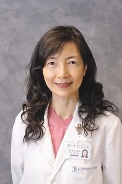 An Asian woman in a white medical coat smiling, with a pink shirt underneath and a name badge, standing against a gray background.