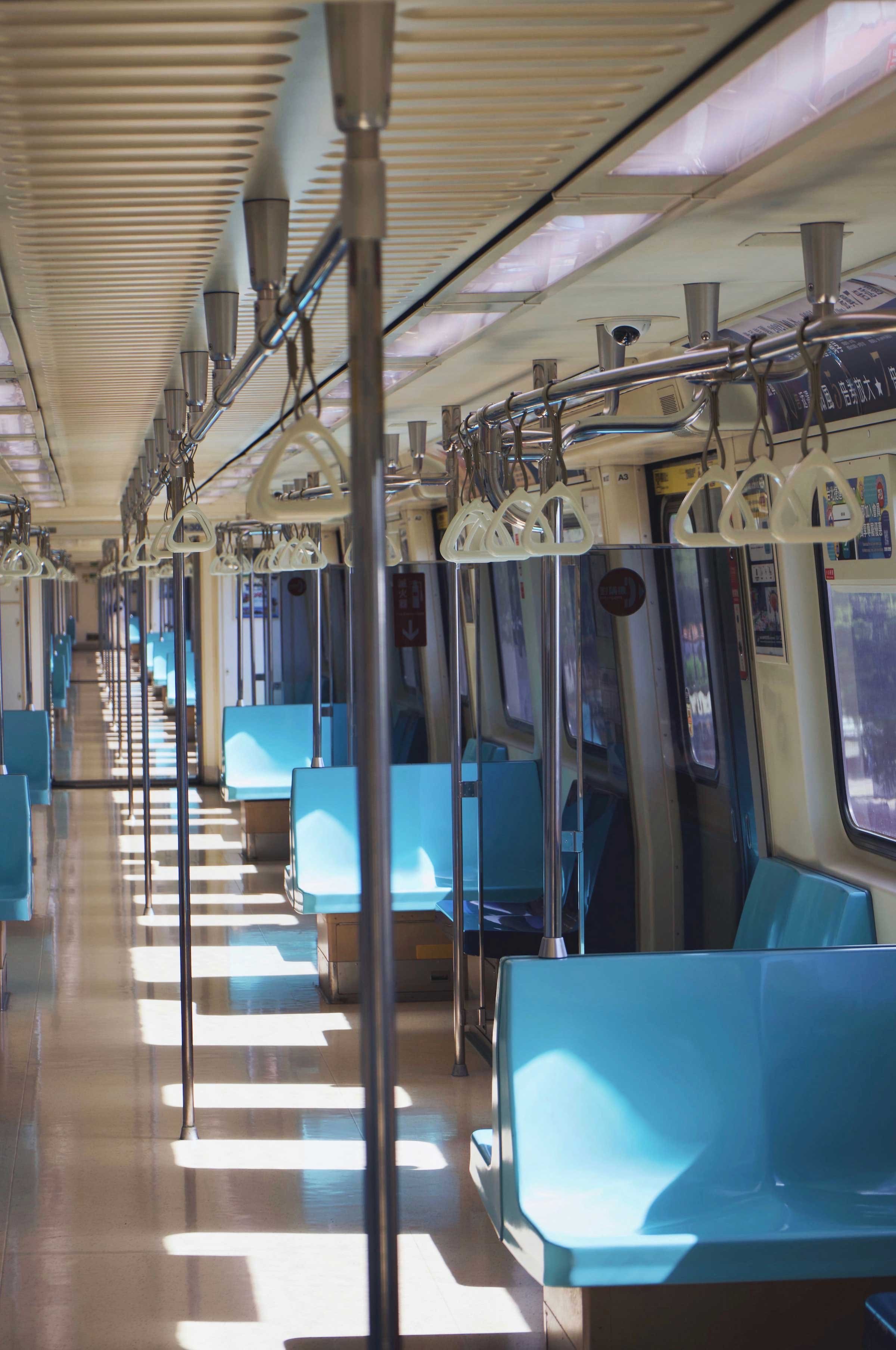 Empty subway train car with blue seats, metal handrails, and hand straps, bathed in natural light from windows.