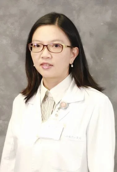 A woman wearing glasses and a white lab coat standing in front of a gray background.