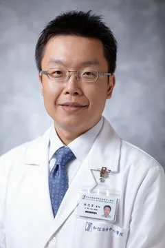 Professional portrait of a male doctor wearing glasses, a white lab coat, and a blue tie, posing against a neutral background.