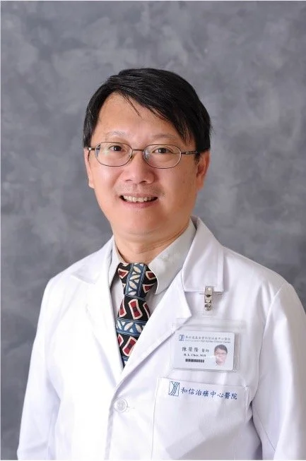 A male doctor wearing glasses, a white lab coat, and a patterned tie, standing in front of a gray background.