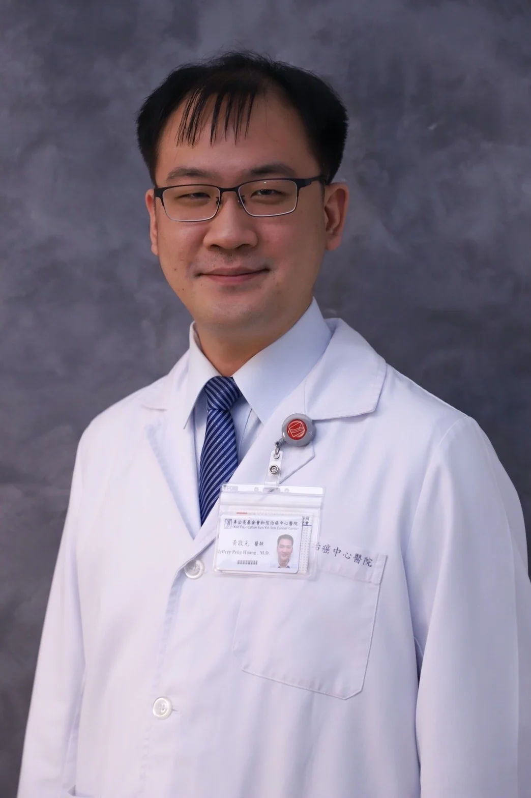 Portrait of a male doctor wearing glasses, a white coat, a blue striped tie, and an ID badge, standing against a gray background.
