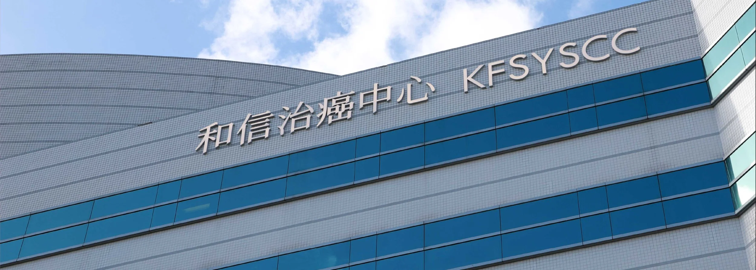The exterior of a modern building with blue glass windows and Japanese characters alongside English text that reads 'KESSCC' against a blue sky with some clouds.