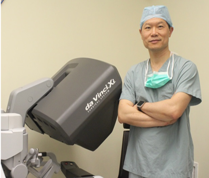 A surgeon in scrubs and a surgical cap standing next to a da Vinci surgical robot.