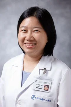 Portrait of a female healthcare professional with black hair, wearing a white lab coat and a name badge, smiling against a gray background.