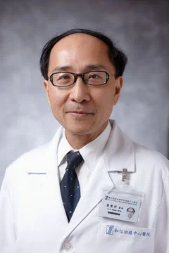 A male doctor wearing glasses, a white coat, and a name badge, standing against a gray background.