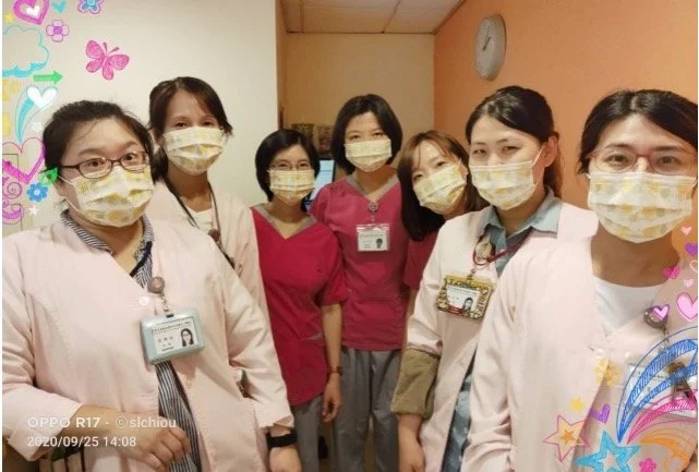 Seven healthcare professionals wearing masks and badges standing together in a hospital corridor, with colorful decorations on the edges of the photo.