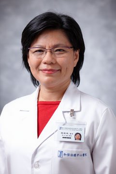 A woman in a white lab coat wearing glasses and a red shirt, smiling at the camera with a gray background.