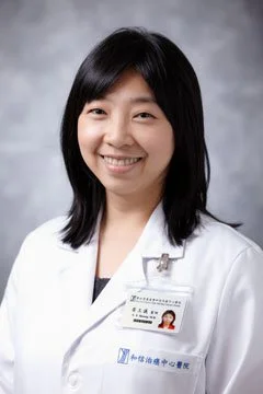 A woman in a white medical coat with a name badge, smiling in front of a gray background.