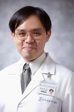 Portrait of a male doctor wearing a white coat and glasses, smiling with a name badge.