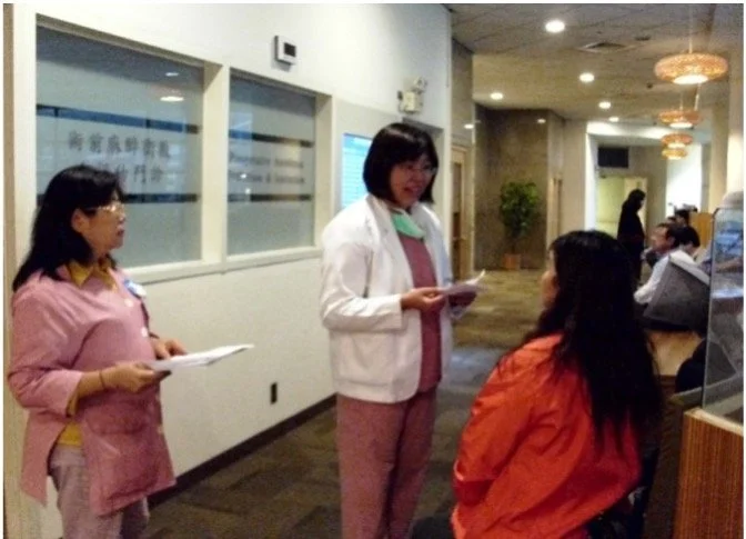 Two women are standing and a woman is sitting in a waiting area of a medical or professional office. The standing women are engaging with each other, while the seated woman is listening. The background shows other people, a water dispenser, and decor