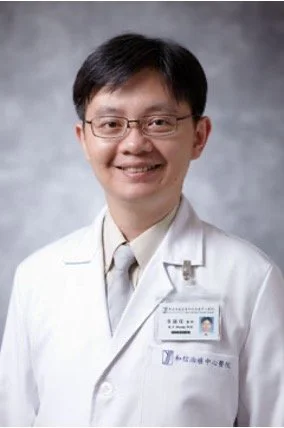 A male doctor wearing glasses and a white lab coat, standing in front of a gray background, smiling for the camera.