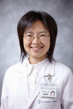 Portrait of a woman with shoulder-length dark hair, glasses, smiling, wearing a white lab coat and professional attire.