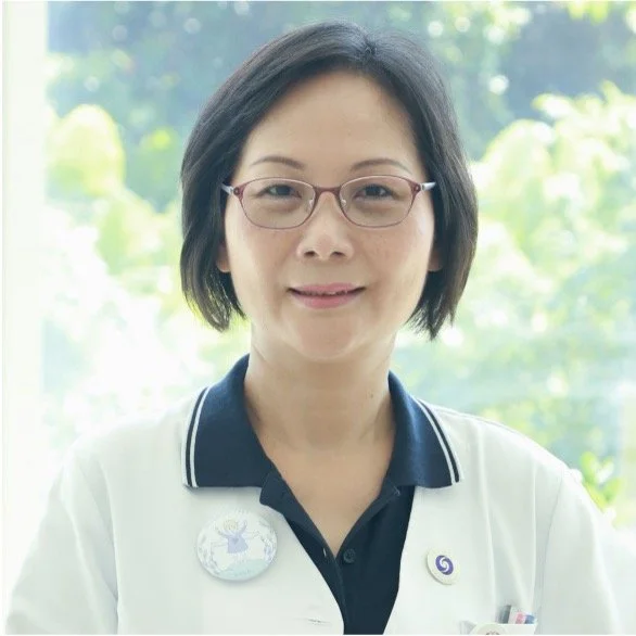 A woman with short black hair and glasses, wearing a white coat over a dark collared shirt, standing outdoors with blurry green trees in the background.