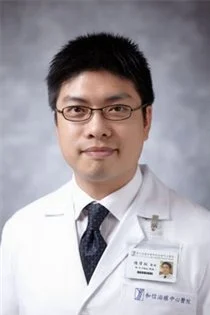 A male doctor with short black hair and glasses, wearing a white coat and a dark tie, posed against a gray background.