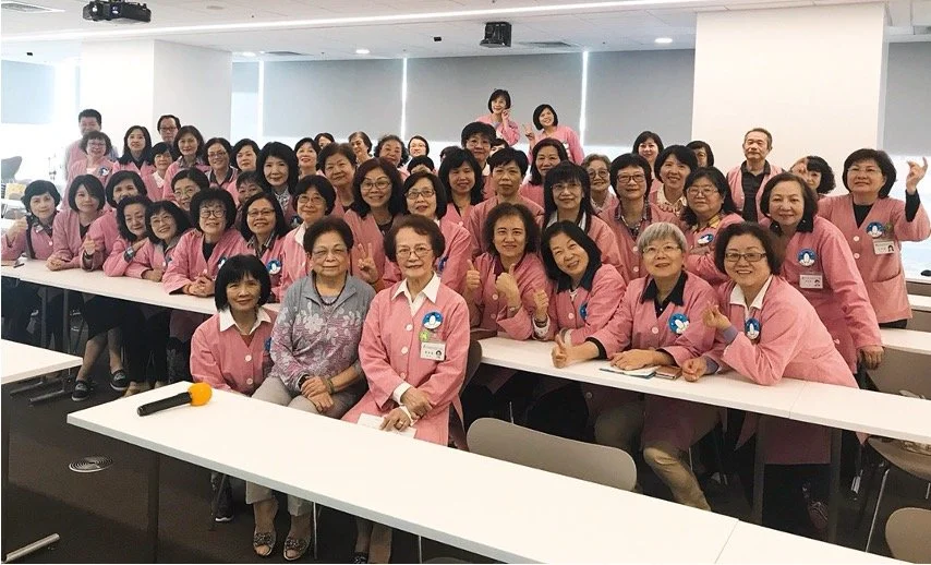 Group of women in pink uniforms gathered in a bright conference room, some sitting and some standing, smiling and making hand gestures for a photo.
