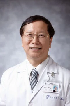 A smiling male doctor wearing glasses and a white lab coat, with a name tag, against a gray background.