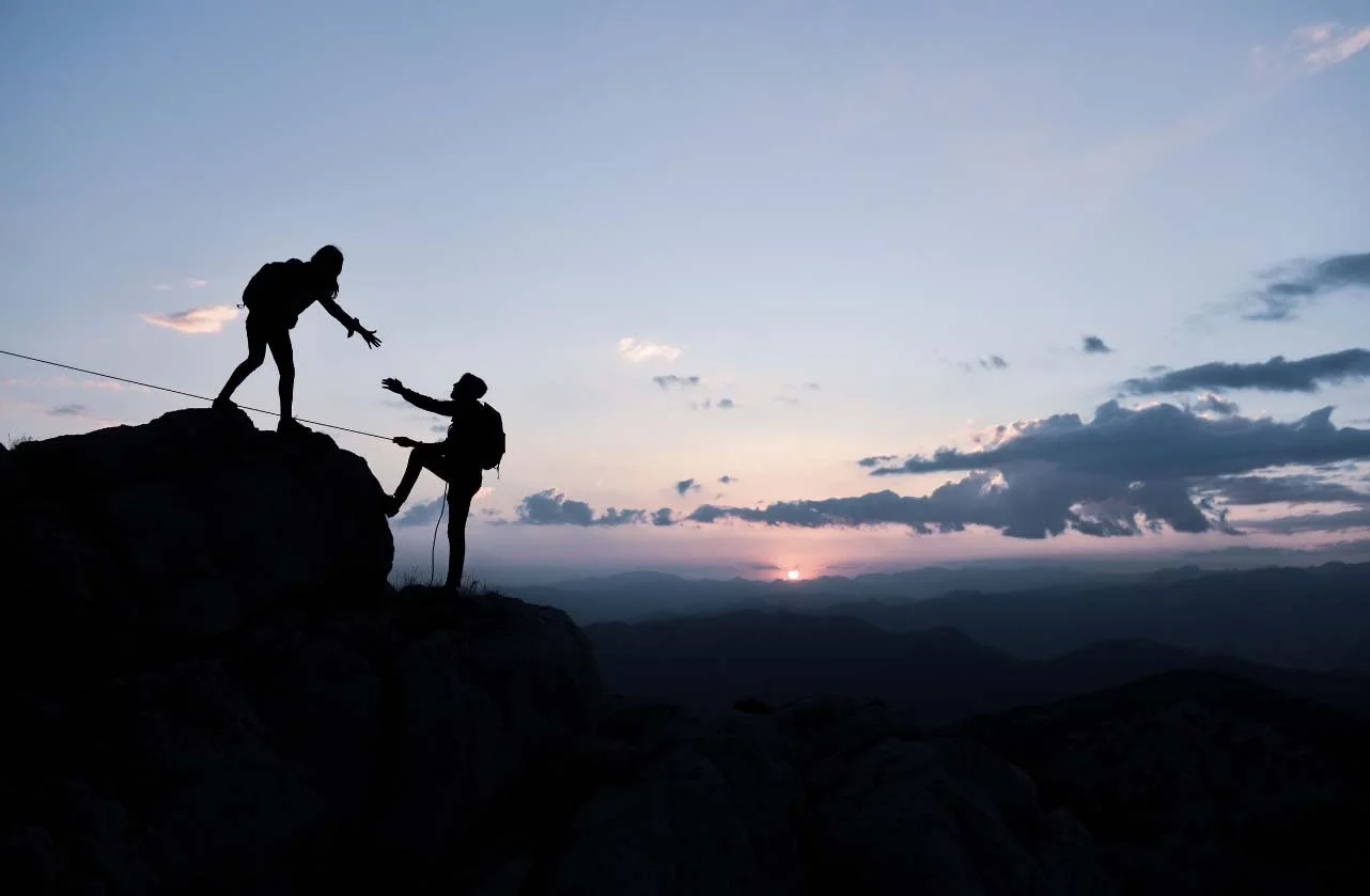Two people silhouetted against a sunset on a mountain, one helping the other climb up a rocky ledge using a safety rope.